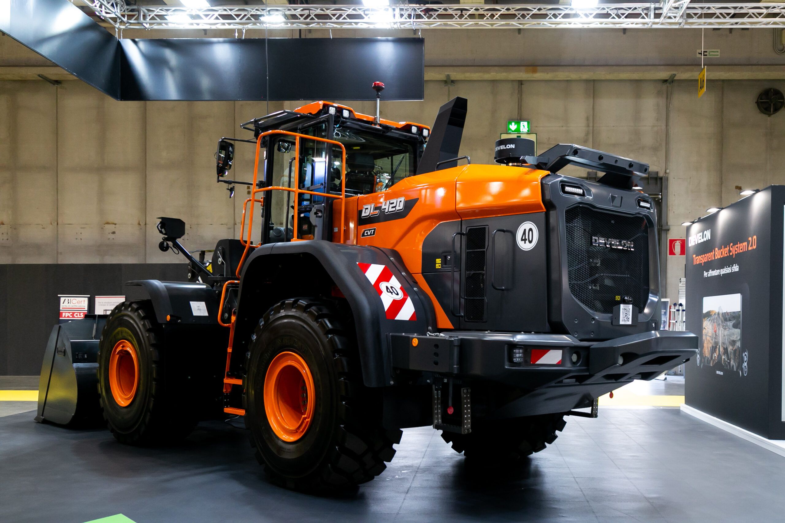 Photograph of a large orange and black Develon DL420CVT-7 wheel loader displayed at an indoor exhibition hall, viewed from the rear three-quarter angle with a 40km/h speed badge visible on the cab