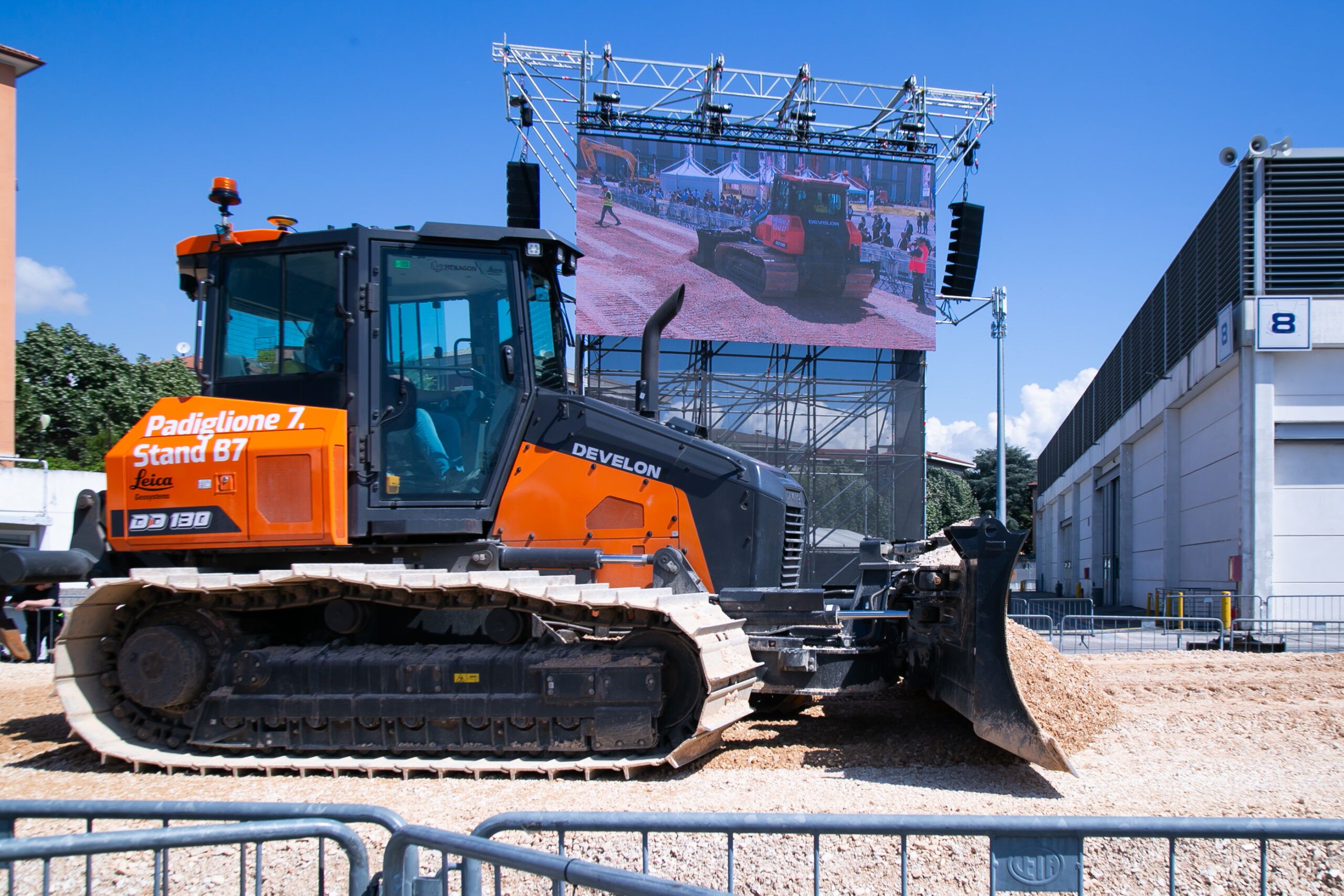 Photograph of an orange and black Develon DD130-7 dozer with Leica Geosystems machine control branding, parked on gravel in an outdoor demonstration area beneath a large display screen.