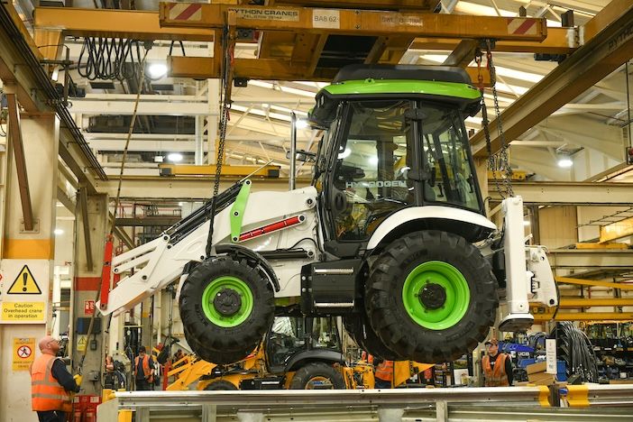 A white and green JCB 3CX Hydrogen backhoe loader suspended by chains from an overhead Python crane on the production line at JCB's Staffordshire factory, with workers in hi-vis jackets visible on the factory floor below