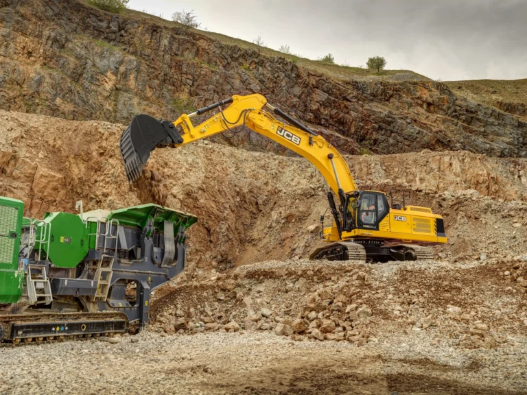 JCB 520X crawler excavator loading excavated material into a green mobile tracked crusher in a quarry