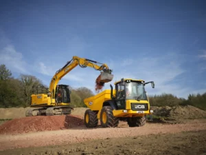 A yellow JCB 715 articulated dump truck being loaded with soil and aggregate by a yellow JCB 150X tracked excavator on a construction site, with woodland and blue sky in the background