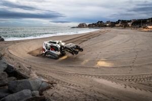 A white and red Bobcat T650 compact track loader with a wide attachment grooming sand on a beach at dusk, with the sea and a coastal town visible in the background