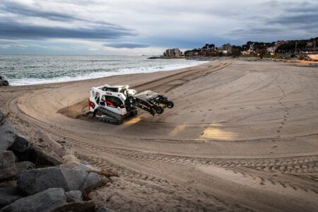 A white and red Bobcat T650 compact track loader with a wide attachment grooming sand on a beach at dusk, with the sea and a coastal town visible in the background