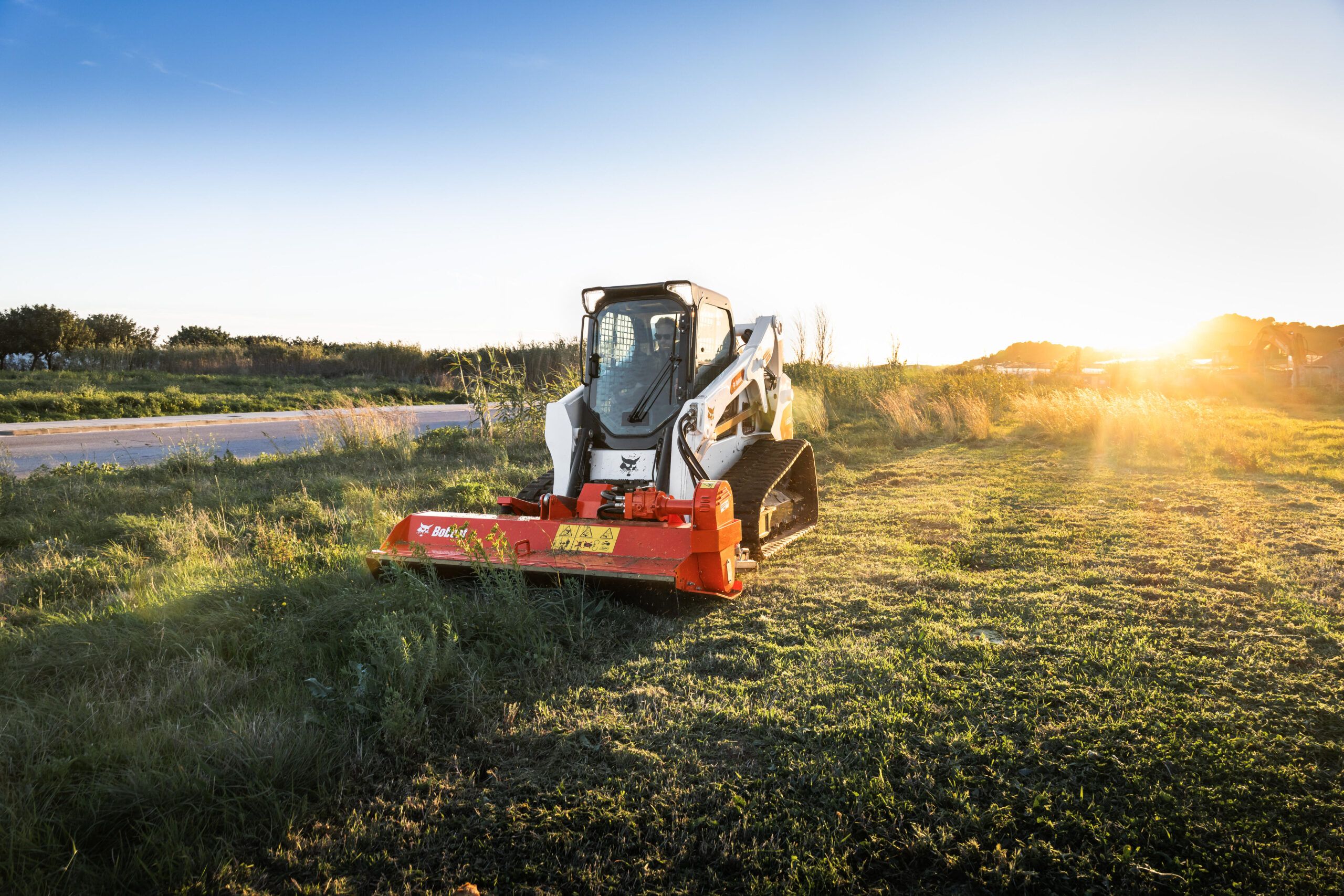 A white Bobcat T650 compact track loader operating a red flail mower attachment in a grass field at sunset, with trees and a road in the background