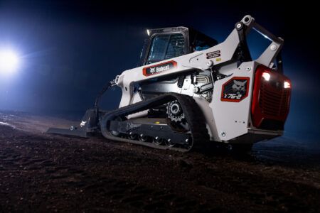 Studio photograph of a white Bobcat T76-2 R-Series compact track loader with red detailing, shown on dark soil under dramatic side lighting against a black background.