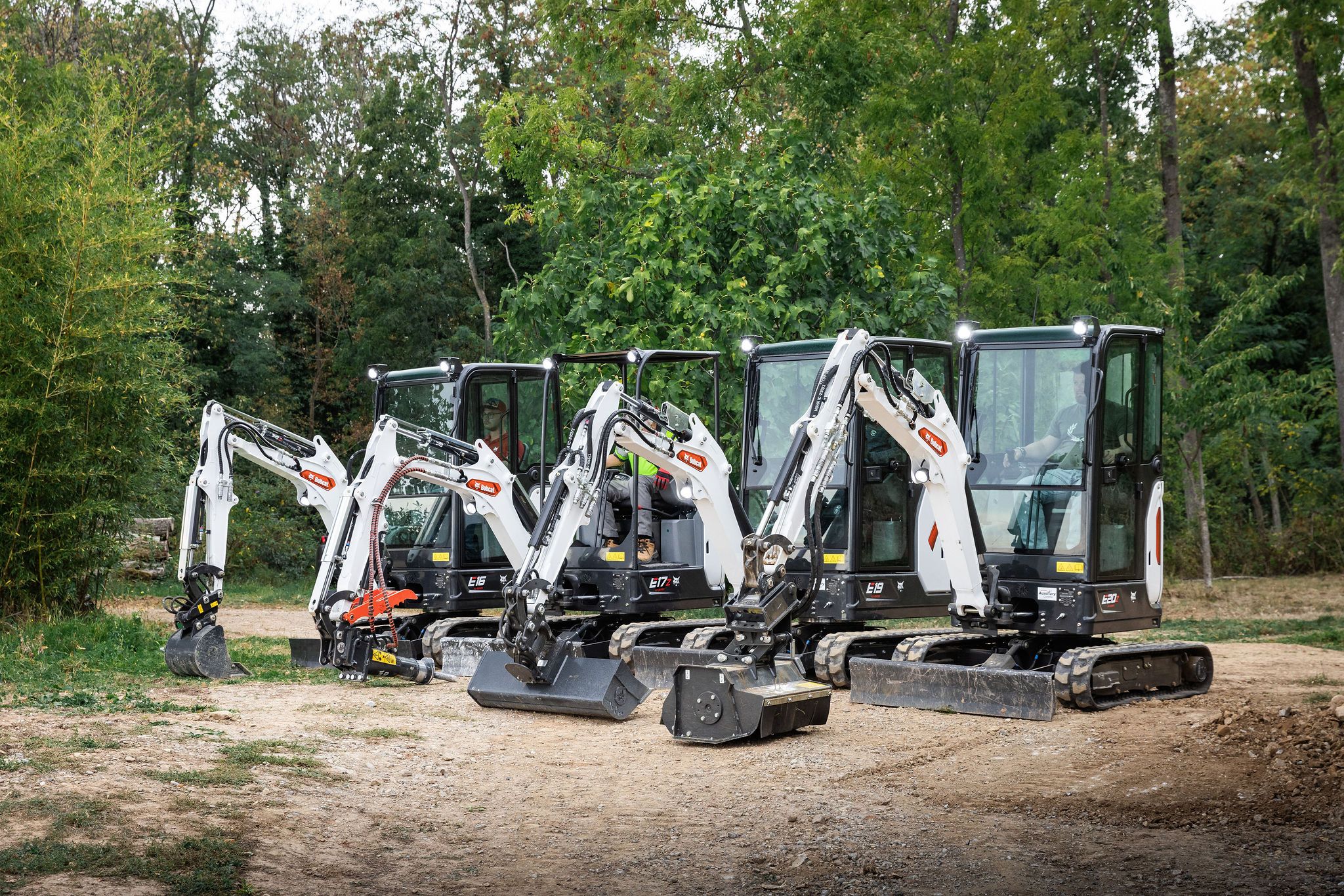 Four Bobcat mini excavators — the E16, E17z, E19 and E20z — lined up on a dirt track in front of dense green woodland, each fitted with a different attachment