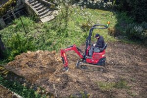 Photograph of a red and black Yanmar SV10 mini excavator working on broken concrete in front of a residential building, with the boom and bucket extended