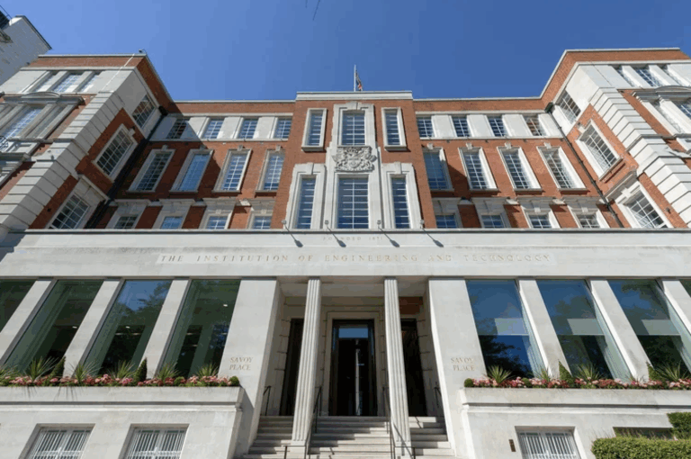 Photograph of the external façade of IET Savoy Place in London, a red-brick and white stone building bearing the inscription 'The Institution of Engineering and Technology', shown from a low angle against a clear blue sky.