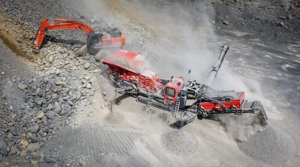 Aerial view of a red Finlay impact crusher operating in a quarry with dust rising around the machine and an orange excavator feeding it from the left