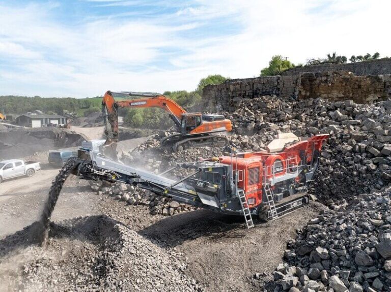 Finlay to debut I-130RS impact crusher at Hillhead 2026 Alt text: Red Finlay J-1280 mobile jaw crusher processing material in a quarry, fed by an orange tracked excavator with broken rock in the foreground and a stone wall behind
