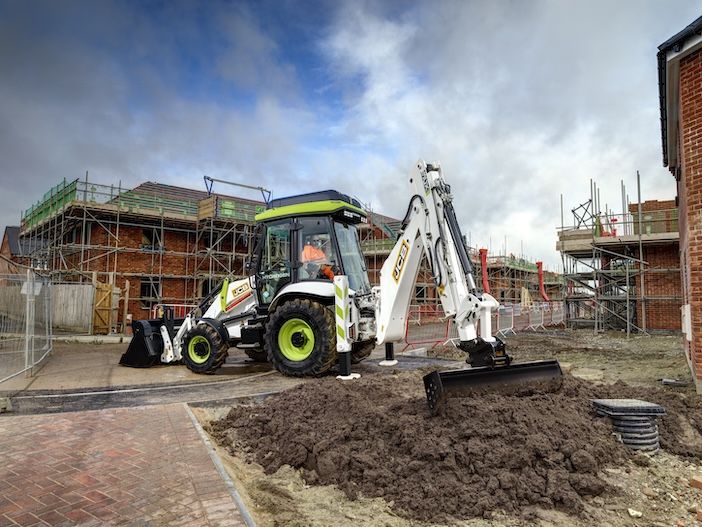 The white and green JCB 3CX Hydrogen backhoe loader working on a residential construction site, with the rear excavator arm fully extended and bucket dug into a mound of earth, scaffolded brick buildings under construction visible in the background under a cloudy sky