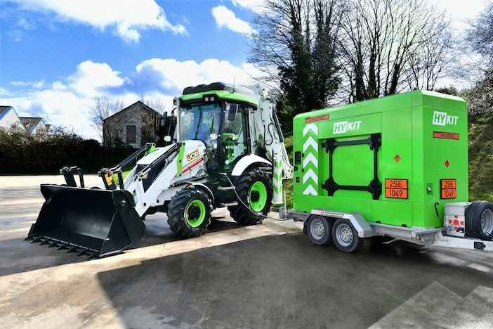 The white and green JCB 3CX Hydrogen backhoe loader, with three hydrogen tanks visible on the cab roof, connected via refuelling hose to the bright green Hykit MHR-X75 trailer-mounted mobile hydrogen refueller, parked on a hardstanding area with trees and a stone building in the background