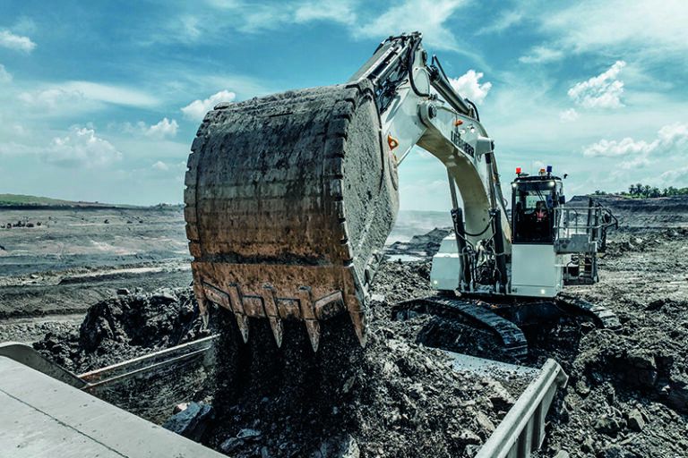 White Liebherr R 9100 mining excavator in backhoe configuration tipping a large bucket of material into a loading area at an open-cast mine