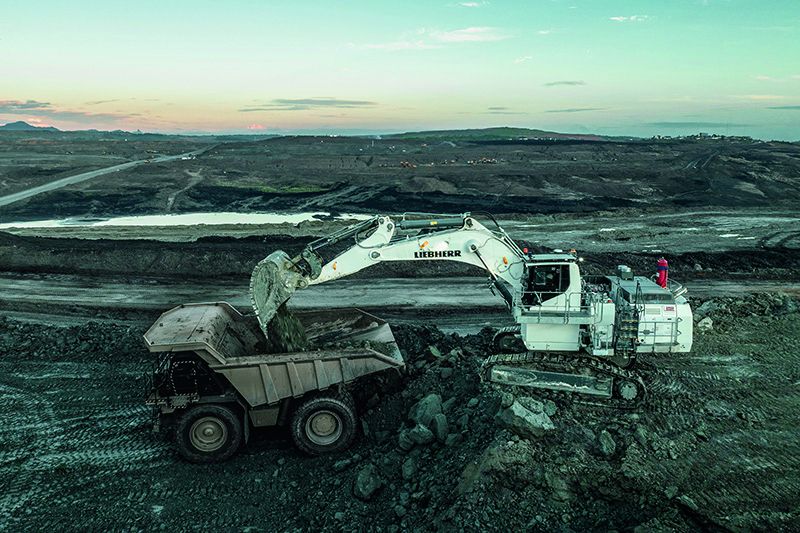 Aerial view of a white Liebherr R 9100 mining excavator loading material into a rigid haul truck at dusk, with an expansive open-cast mine in the background