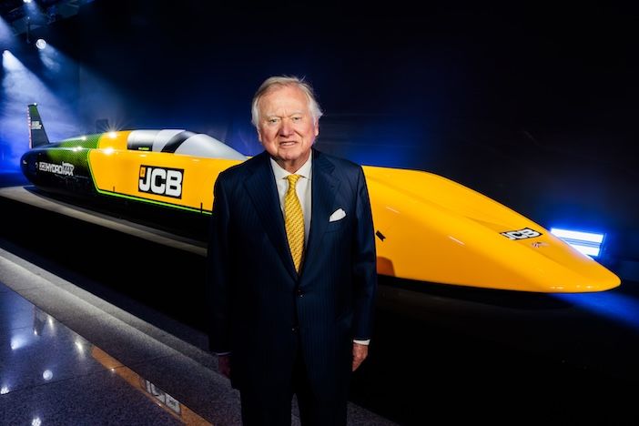 JCB chairman Lord Bamford, dressed in a dark suit with a yellow tie, stands in front of the full-length JCB Hydromax land speed record car, a low-slung yellow and green streamliner with JCB branding, displayed under dramatic stage lighting at a reveal event
