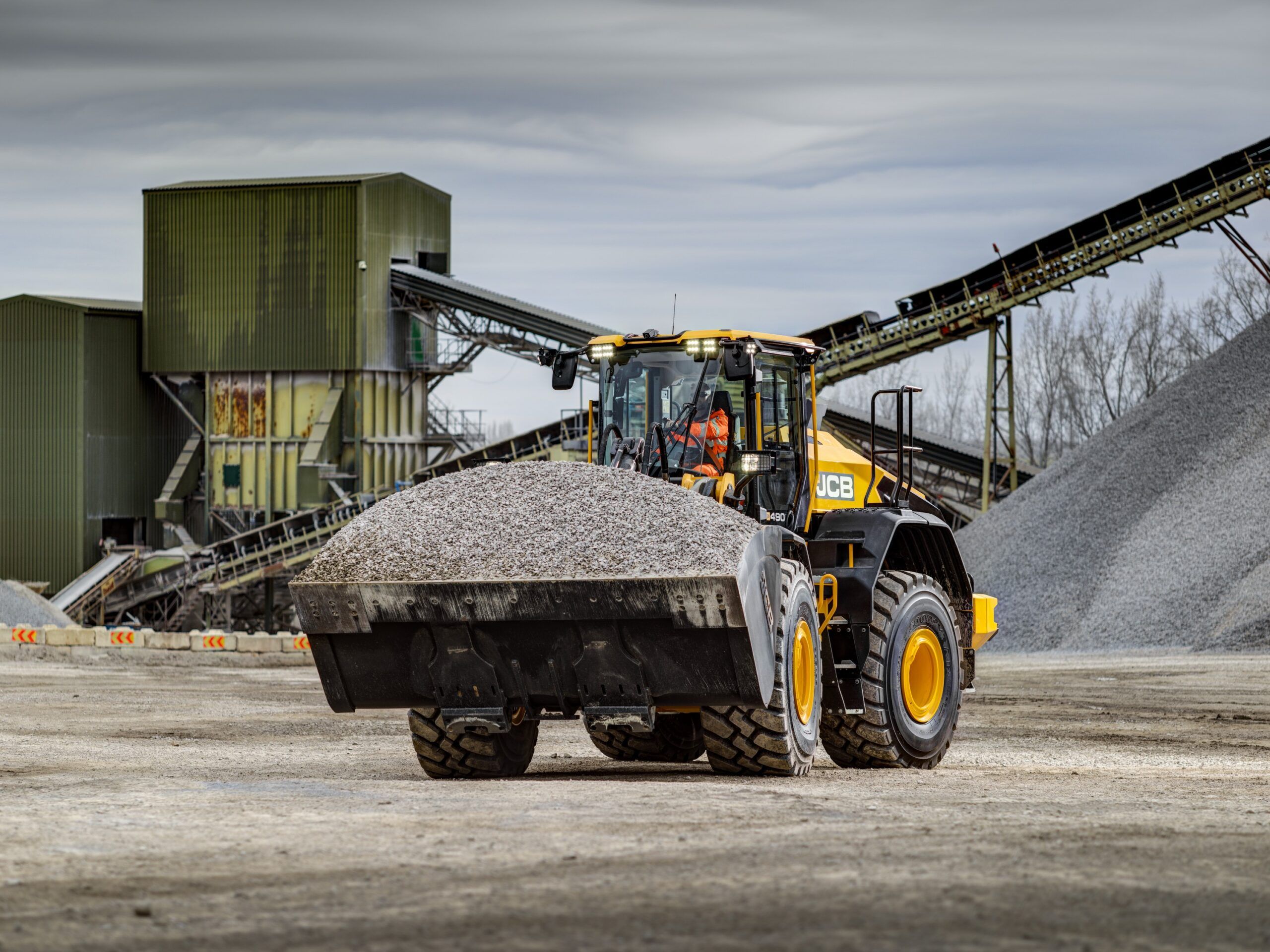 JCB 490 wheeled loader with a full aggregate bucket at a quarry site, with conveyor infrastructure visible in the background