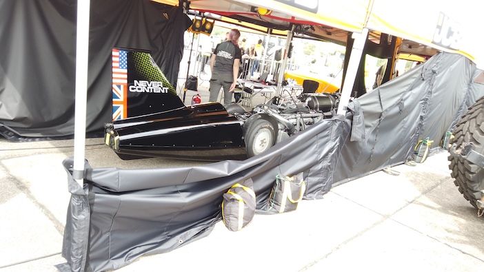  The JCB Hydromax partially covered with black sheeting in a pit area, with the "Never Content" tail fin and US and UK flag livery visible at the rear