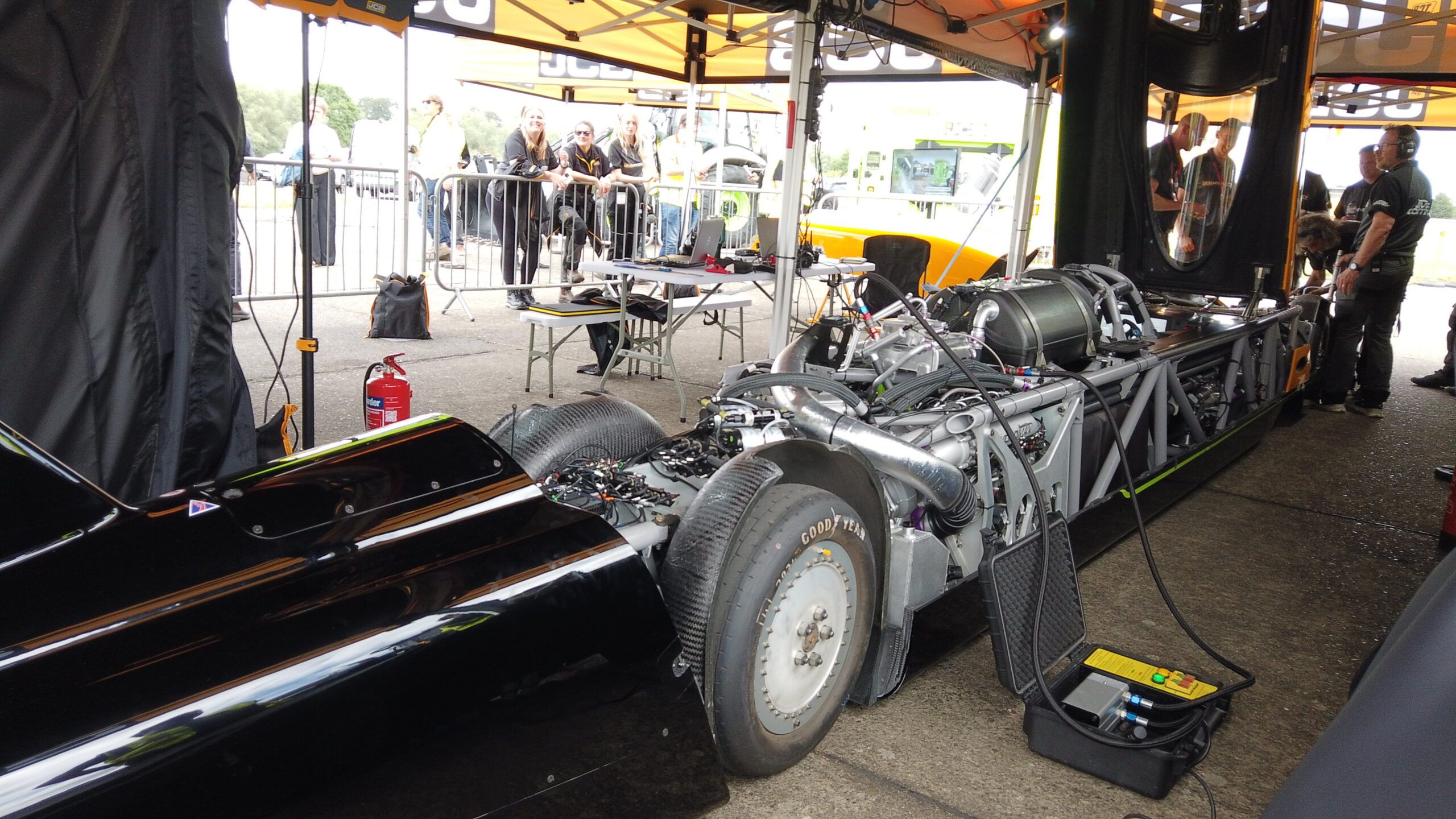 Exposed chassis of the JCB Hydromax under a JCB awning, showing engine, turbocharging and wiring, with crew and spectators behind
