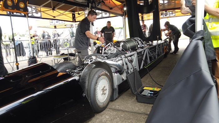 A mechanic in a black JCB team shirt works on the exposed drivetrain and Goodyear tyres of the JCB Hydromax, with other crew members visible in the background under a team canopy