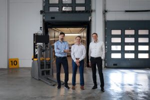 Three men stand beside an autonomous pallet truck at an open loading dock door, with a trailer in the background