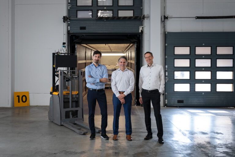 Three men stand beside an autonomous pallet truck at an open loading dock door, with a trailer in the background