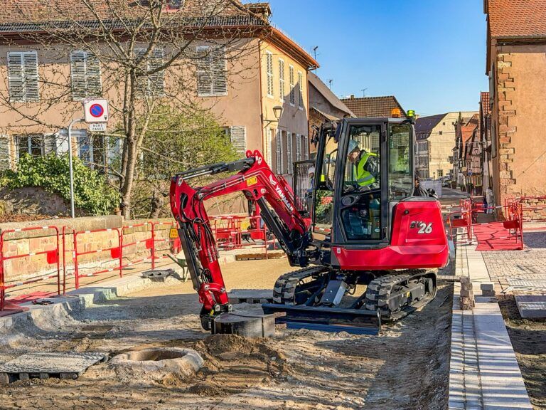 Red Yanmar SV26 mini excavator operated by a worker in high-visibility clothing on an urban street construction site, with buildings and safety barriers visible in the background