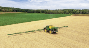 An aerial view of a green and yellow John Deere 500R self-propelled sprayer working across a bare field with its boom fully extended, alongside a green crop field and tree line.