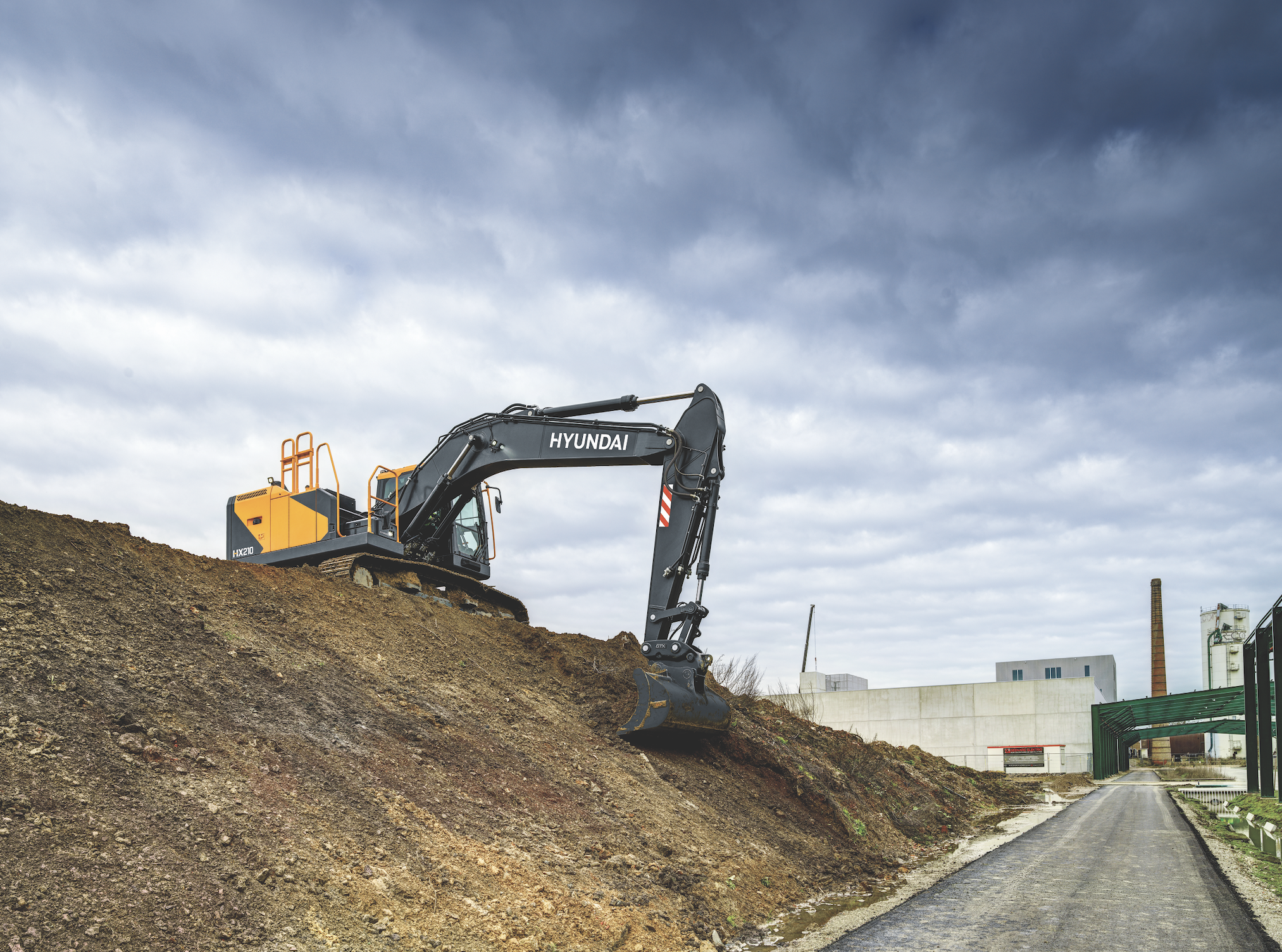 Hyundai HX210L Next Generation crawler excavator operating on an earthworks slope with an industrial building in the background