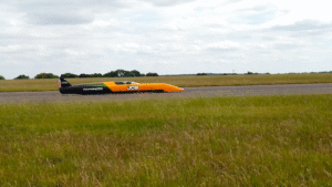 A low-slung orange and black JCB Hydromax land speed record car running along a flat runway, with open grassland and trees in the background Caption: The JCB Hydromax during shakedown runs at RAF Wittering in Cambridgeshire