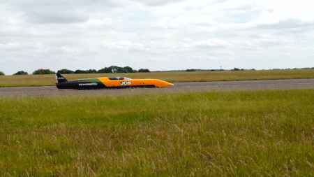 A low-slung orange and black JCB Hydromax land speed record car running along a flat runway, with open grassland and trees in the background Caption: The JCB Hydromax during shakedown runs at RAF Wittering in Cambridgeshire
