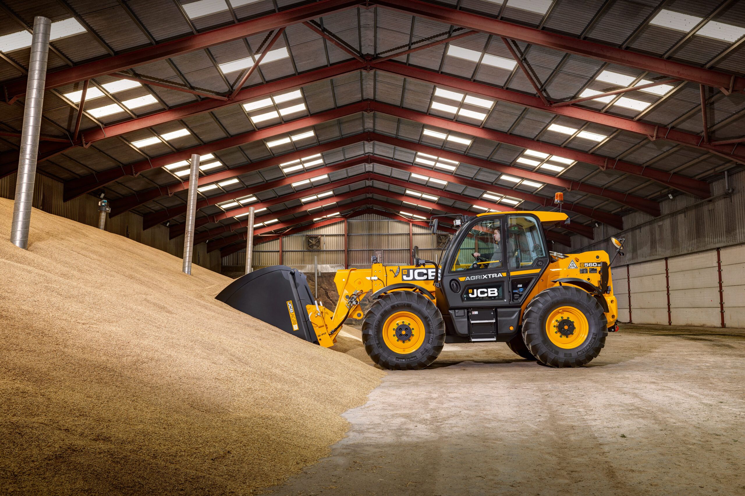 A yellow JCB 560-80 AGRIXTRA telehandler loading grain from a large heap inside a farm storage building, with its bucket lowered into the pile