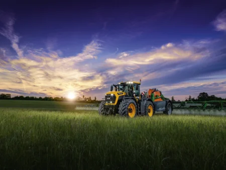 New JCB Fastrac 6300 to lead Cereals lineup A yellow JCB Fastrac 6300 tractor towing a green Amazone trailed sprayer with extended booms across a field at sunset, applying spray to a standing crop