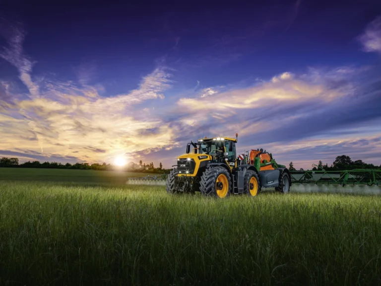 New JCB Fastrac 6300 to lead Cereals lineup A yellow JCB Fastrac 6300 tractor towing a green Amazone trailed sprayer with extended booms across a field at sunset, applying spray to a standing crop