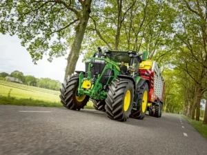John Deere 6R large frame tractor towing a red and yellow trailed implement along a tree-lined road