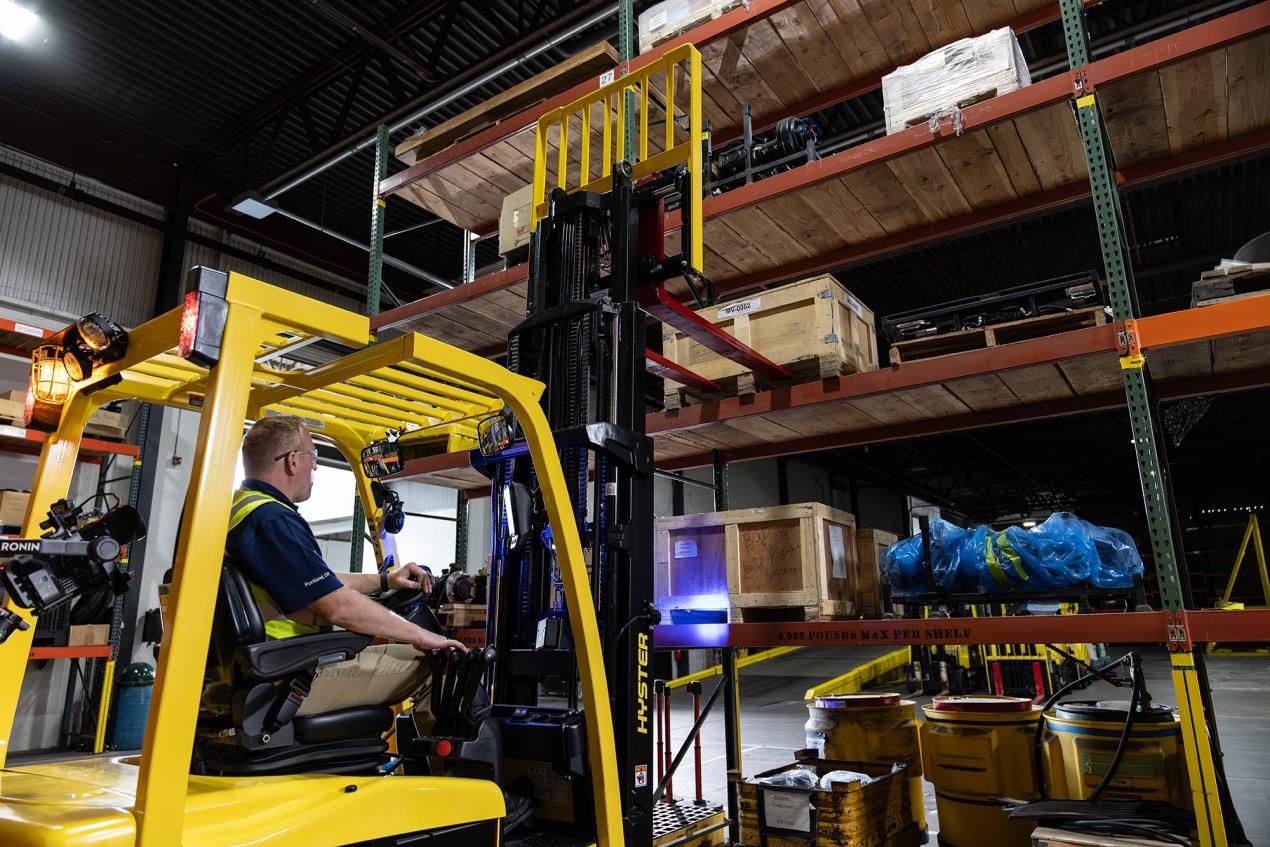 Operator in a yellow Hyster XN2 electric forklift raising the mast in a parts warehouse, with high-racking shelving and crated components visible in the background.