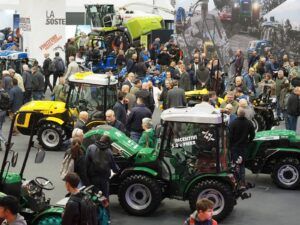 Busy indoor exhibition hall at EIMA with yellow and green compact tractors and visitors browsing stands