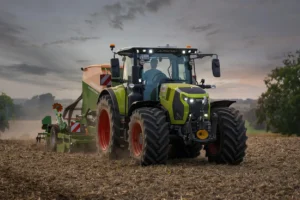 A green Claas Arion tractor working a stubble field at dusk with its headlights on, towing a seed drill