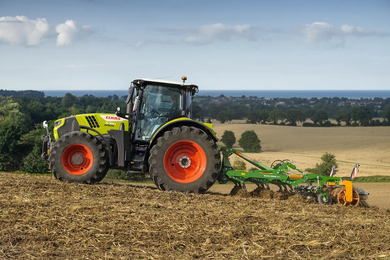 A Claas Arion 6180 tractor in profile working a hillside field, with a coastal landscape in the background.