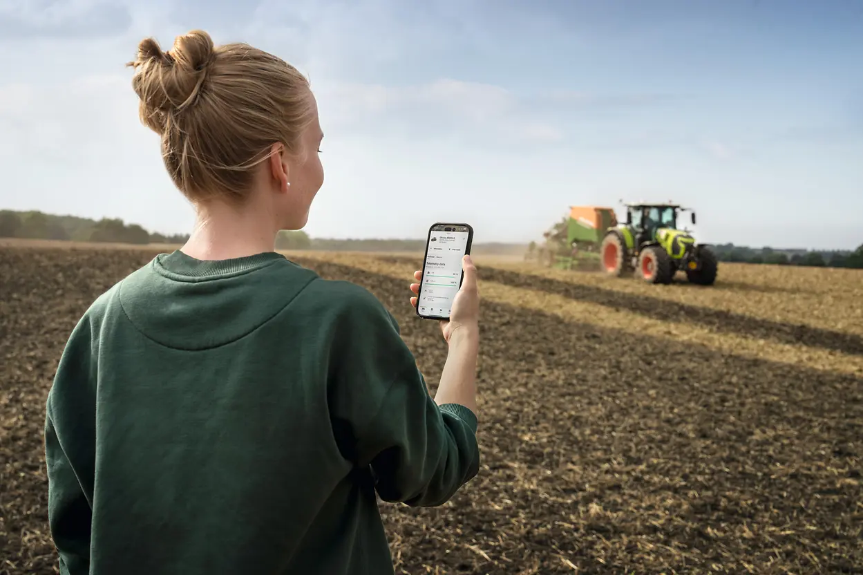 A person seen from behind holding a smartphone displaying telemetry data, watching a Claas Arion tractor work a field.