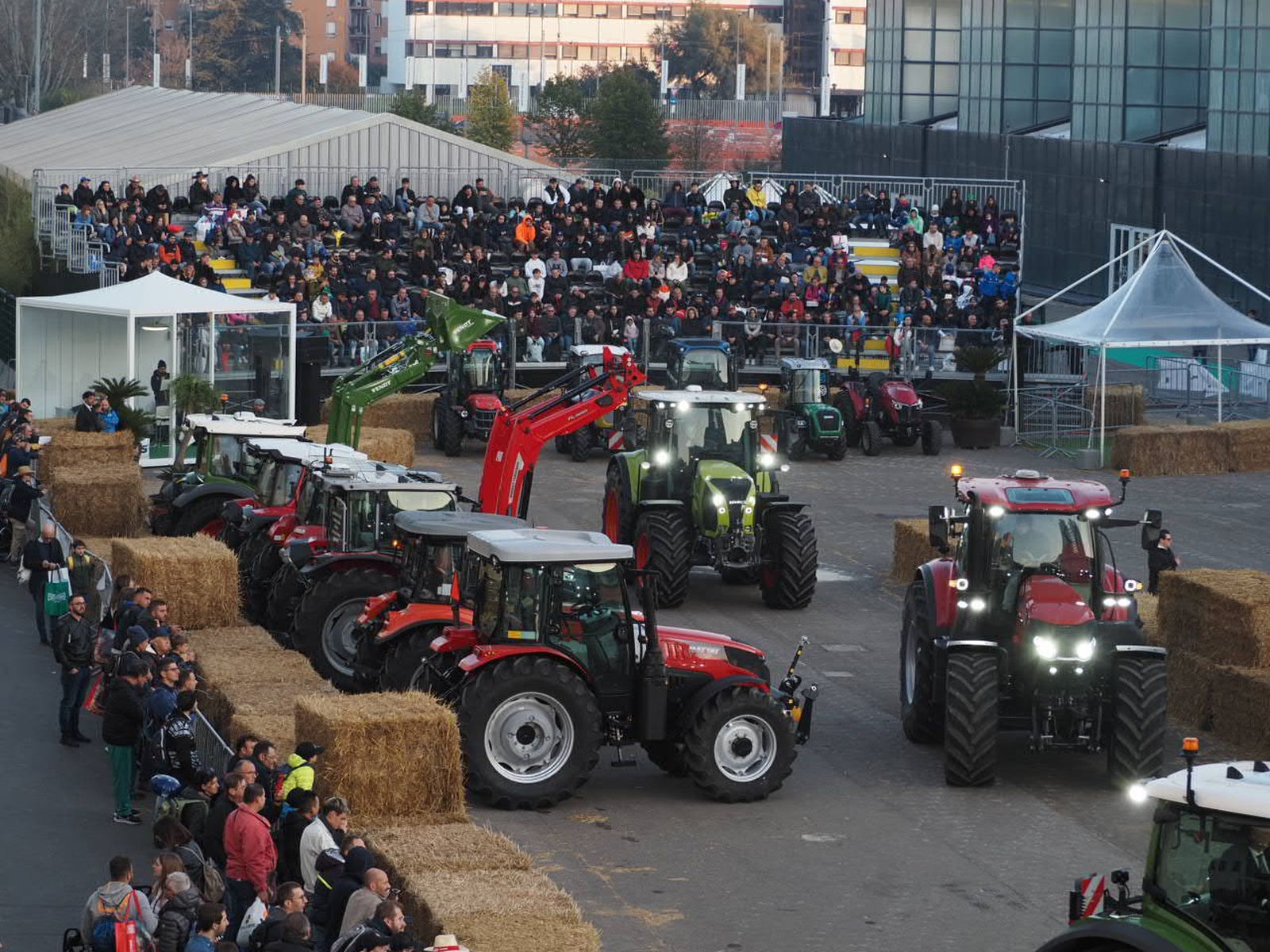 Row of tractors on an outdoor course lined with hay bales, watched by a large seated crowd at dusk