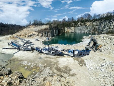 Kleemann to bring crushing and screening lineup to Steinexpo A line of Kleemann crushing and screening plants working in a quarry beside a turquoise water pool, fed by a tracked excavator