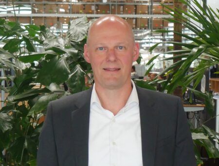 Linak names Michael Jensen president of US business Michael Jensen, wearing a dark suit and open-collar white shirt, standing in front of greenery in a production facility
