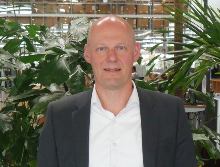 Linak names Michael Jensen president of US business Michael Jensen, wearing a dark suit and open-collar white shirt, standing in front of greenery in a production facility