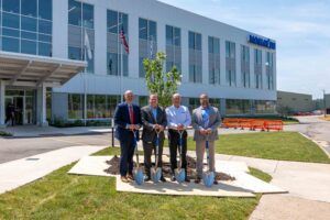 Four men in business attire hold ceremonial spades around a newly planted tree outside Komatsu’s new office building in Peoria, Illinois