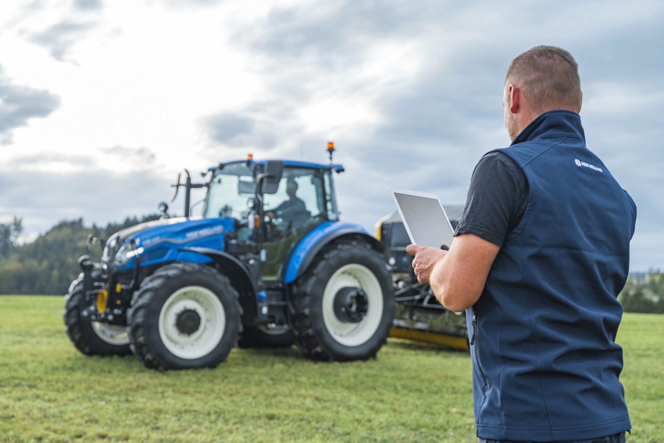A New Holland technician holds a tablet in a field while a blue New Holland T5 tractor works behind him under a cloudy sky