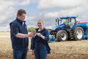 Two New Holland representatives look at a smartphone in a field, with a blue New Holland T7 tractor and implement behind them