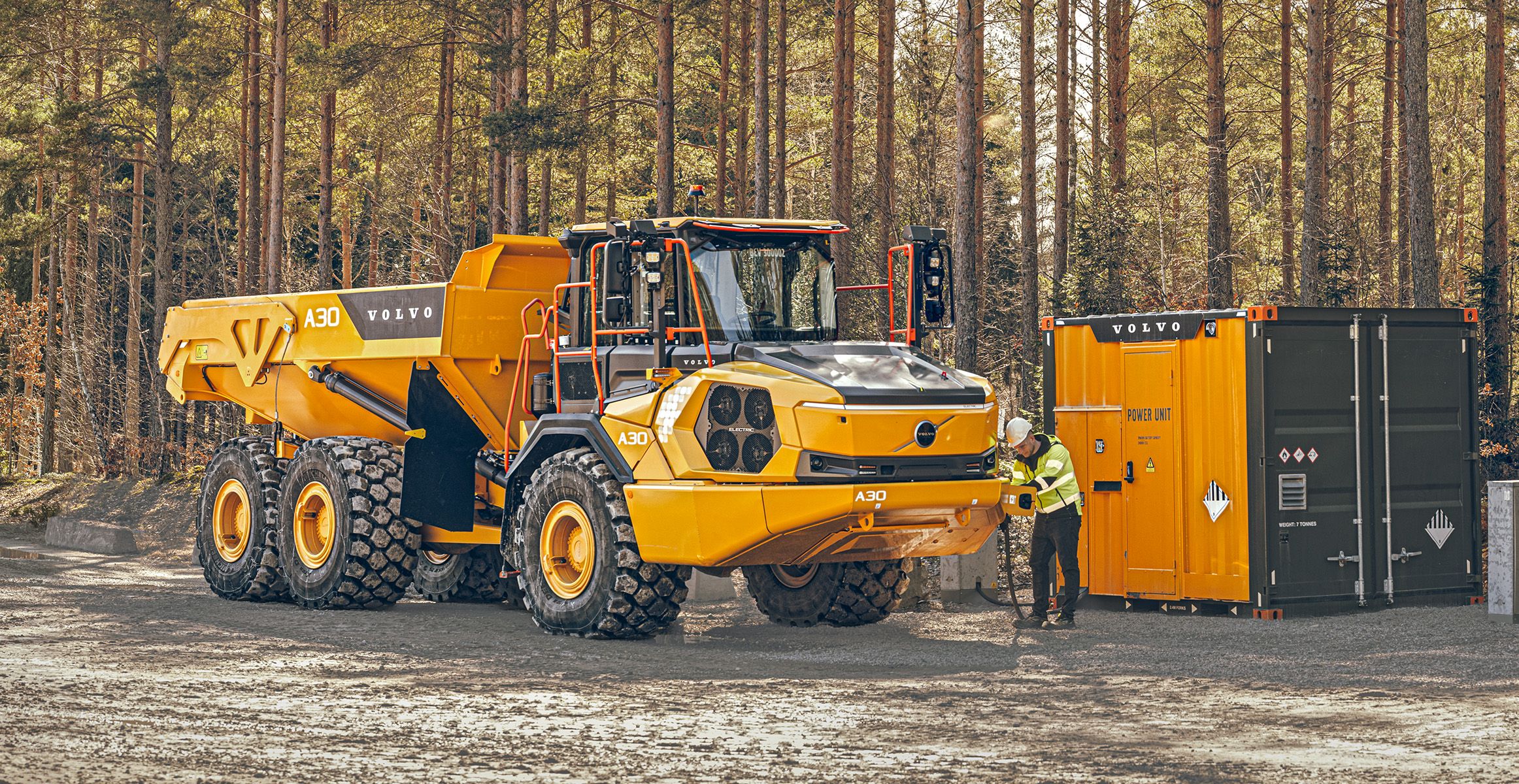 A yellow Volvo A30 Electric articulated hauler parked in a pine forest, connected by cable to an orange Volvo power unit charging container