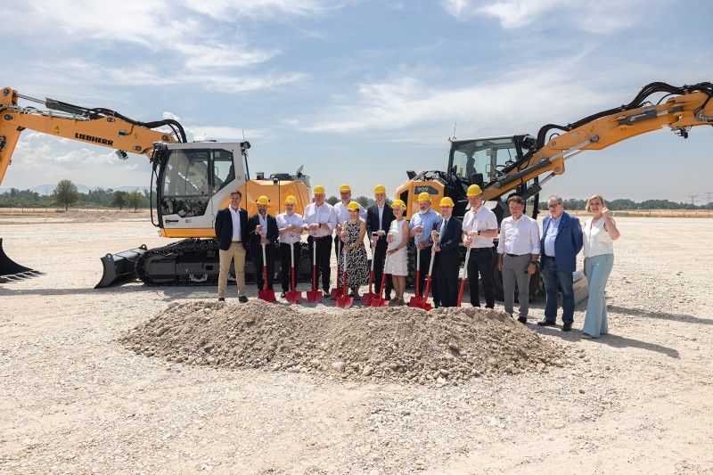 Group in hard hats holding red ceremonial spades at a mound of earth, flanked by two yellow Liebherr earthmoving machines