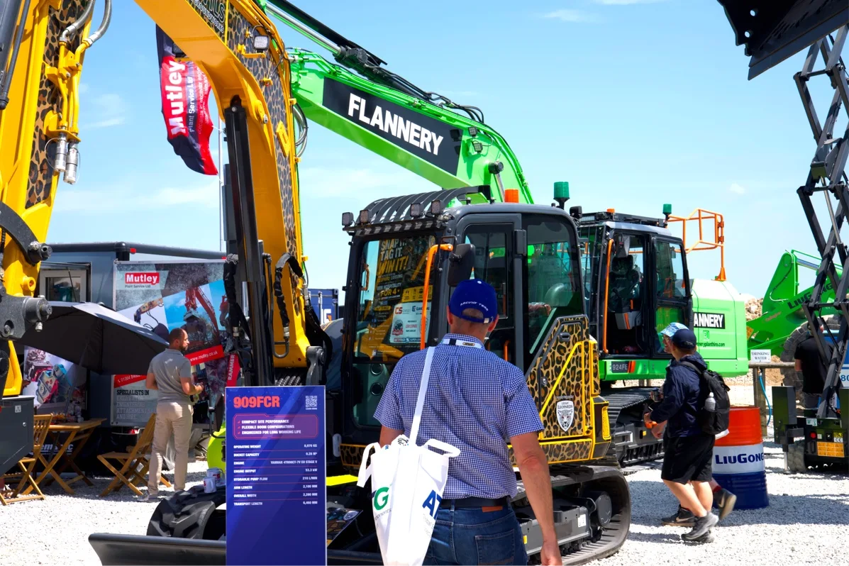 Visitors inspect a LiuGong compact excavator in leopard-print livery, with a spec board in the foreground and a green machine behind.