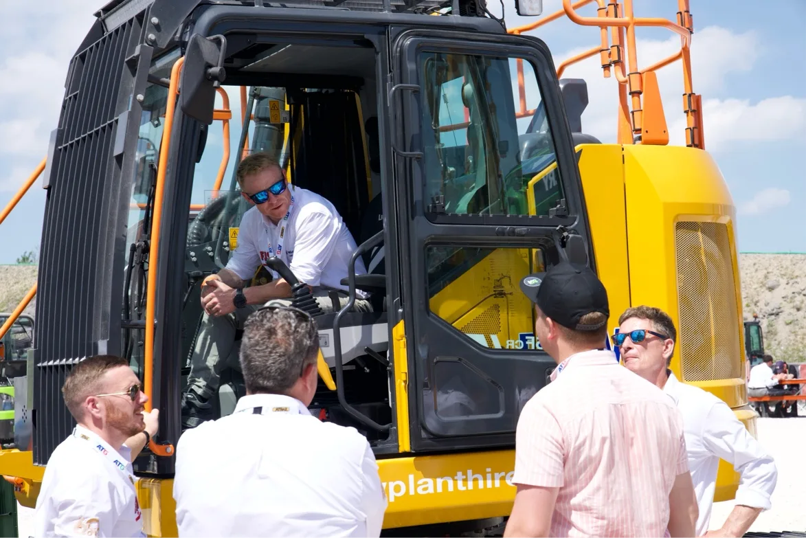 A man sits in the cab of a yellow LiuGong excavator talking with a group of visitors gathered around the machine at the show.
